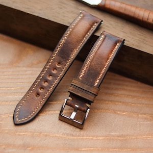 Pode incluir: Duas pulseiras de relógio de couro castanho com costuras claras e fivela prateada. As pulseiras são envelhecidas, mostrando variações de cor. Uma faca e um relógio de bolso também são visíveis.