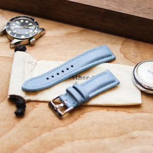 Könnte beinhalten: Hellblaues Leder-Uhrenarmband mit silberner Schnalle, auf einem cremefarbenen Beutel. Eine Vintage-Uhr und eine Stoppuhr sind ebenfalls zu sehen. Die Gegenstände liegen auf einer Holzoberfläche, was auf das Thema Zeitmessung und Accessoires hindeutet.