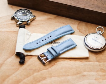 Peut inclure: Un bracelet de montre en cuir bleu clair avec une boucle en métal argenté, posé sur une petite pochette en tissu crème. Le bracelet présente des coutures visibles et plusieurs trous de réglage, près d'une montre-bracelet et d'une montre à gousset.