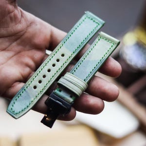 Può includere: Un paio di cinturini per orologio con motivo mimetico verde chiaro e blu. I cinturini in pelle hanno cuciture verdi. Un cinturino ha fori di regolazione, l'altro una fibbia. I cinturini sono tenuti in una mano.