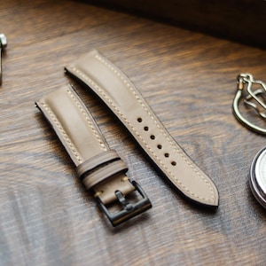 Pode incluir: Um par de pulseiras de relógio em couro castanho claro com costuras contrastantes e uma fivela de metal escuro. As pulseiras são exibidas em uma superfície de madeira, juntamente com um relógio e um cronômetro.