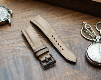 May include: A two-piece taupe leather watch strap featuring light-colored stitching, a black metal buckle, and two matching leather keepers. The strap is positioned between a black-faced wristwatch and a silver-toned pocket watch.