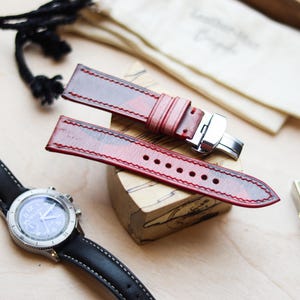 Könnte beinhalten: Rote und braune Leder-Uhrenarmbänder mit Edelstahlverschluss. Eine schwarze Uhr mit silberner Lünette und blauem Zifferblatt ist ebenfalls zu sehen. Die Armbänder liegen auf einem Holzblock.