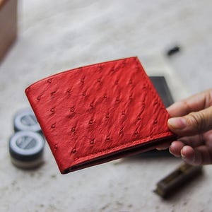 Könnte beinhalten: Ein leuchtend rotes Bifold-Portemonnaie mit einer ausgeprägten strukturierten Oberfläche mit kleinen, gleichmäßig verteilten Federfollikel-Vertiefungen, die für Straußenleder charakteristisch sind. Das Portemonnaie hat gerade Nähte.