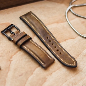 Pode incluir: Pulseira de relógio de couro marrom de duas peças com costura branca e fivela preta. A pulseira é desgastada, mostrando desgaste. A pulseira do relógio está em uma superfície de madeira clara.