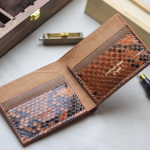 以下が含まれることがあります： 茶色、オレンジ、黒の色合いのヘビ革模様のオープンレザーウォレット。ウォレットは開いており、カードスロットと請求書コンパートメントが表示されています。「Leather Man Design」の文字が内側に表示されています。