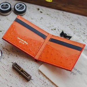 May include: A bi-fold wallet crafted from bright orange textured leather with dark blue interior card slots. The interior features the gold-stamped text Leather-Man Bespoke. The wallet is open, displaying multiple slots for cards and currency.