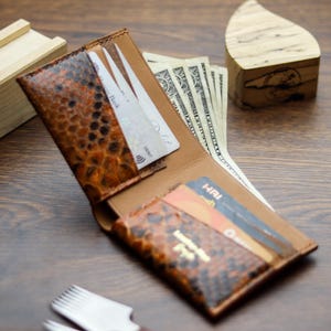 May include: A bifold wallet featuring a textured, brown and black snakeskin-patterned exterior with a tan leather interior. The wallet contains multiple card slots holding various credit cards and a compartment filled with several US dollar bills.