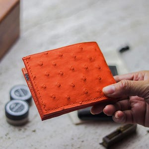 Puede incluir: Una vibrante cartera de cuero naranja con un patrón texturizado distintivo. La cartera se sostiene en una mano, mostrando su forma cuadrada y costuras visibles. La cartera parece nueva y sin usar.