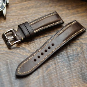 Pode incluir: Close-up de uma pulseira de relógio de couro castanho com costuras creme. A pulseira tem uma fivela prateada e vários orifícios para ajuste. A pulseira do relógio está sobre uma superfície de madeira.