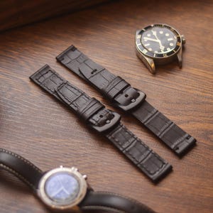 Könnte beinhalten: Zwei schwarze Uhrenarmbänder mit Krokodilmuster und schwarzen Schnallen werden auf einer Holzoberfläche präsentiert. Eine Armbanduhr mit schwarzem Zifferblatt und goldfarbenem Gehäuse ist ebenfalls sichtbar. Die Szene deutet auf einen Fokus auf Uhrenzubehör und Design hin.