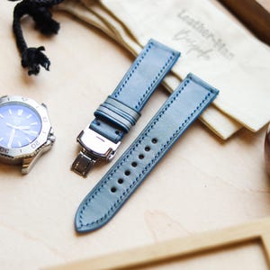 Könnte beinhalten: Ein hellblaues Lederarmband mit weißen Nähten und einer silbernen Schließe. Das Armband liegt neben einer silbernen Uhr mit blauem Zifferblatt. Ein kleiner Beutel mit dem Text "Leather-Man Bespoke" ist ebenfalls im Bild.