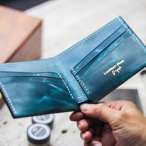 May include: Open teal leather wallet with white stitching. The wallet has multiple card slots and a bill compartment. The text "Leather-Man Design" is printed in gold on the inside. The wallet is open, revealing the interior.