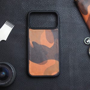 Può includere: Custodia per telefono nera con motivo mimetico nei toni del marrone e dell'arancione. La custodia ha una cornice nera. Altri elementi nell'immagine includono un cronometro, un portafoglio, un obiettivo della fotocamera e strumenti in pelle.