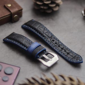 Pode incluir: Duas pulseiras de relógio de couro preto com costuras azuis. Uma pulseira tem uma fivela de metal e a outra tem uma borda azul.