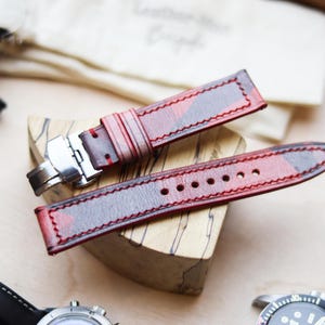 Può includere: Cinturini per orologi in pelle rossa e nera con chiusura in acciaio inossidabile. I cinturini sono esposti su un blocco di legno. I cinturini hanno cuciture rosse e un motivo. Un orologio con cinturino nero è visibile sullo sfondo.