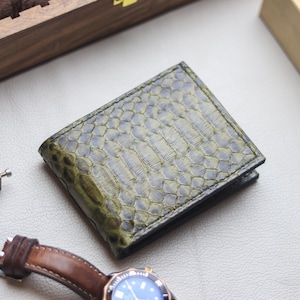 May include: A bi-fold wallet featuring a textured, olive green snakeskin pattern on the exterior. The wallet has a rectangular shape with visible scale details across the surface, showcasing a gradient of dark and light green tones throughout.
