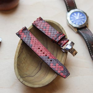 Könnte beinhalten: Rote und graue Schlangenleder-Uhrenarmbänder mit Edelstahlverschluss, ausgestellt in einer Holzschale. Ein braunes Leder-Uhrenarmband und eine Uhr mit blauem Zifferblatt sind ebenfalls zu sehen.