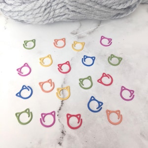 May include: A selection of cat-shaped stitch markers in a range of colours, such as green, pink, yellow, orange, and blue. The markers are scattered on a white surface, with a ball of grey yarn in the background. Suitable for knitting and crochet.