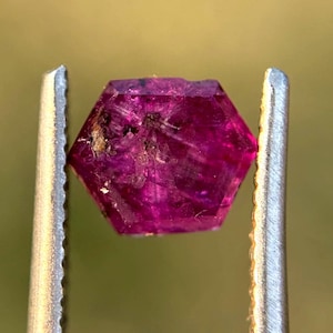 May include: A faceted, hexagonal ruby gemstone held by silver tweezers. The ruby is a deep, saturated pink-red color with visible internal inclusions. The tweezers are in sharp focus, highlighting the gem's clarity and shape.