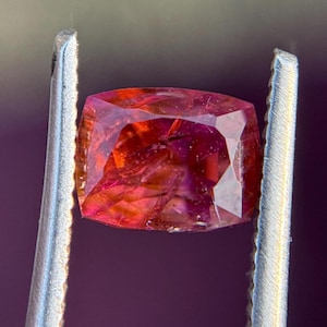 May include: A faceted, reddish-orange gemstone held by silver tweezers. The gem has a cushion cut with visible internal inclusions and a vibrant, translucent appearance. The background is a blurred, dark purple.