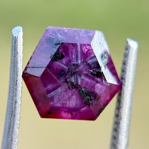 May include: A faceted, hexagonal ruby gemstone held by metal tweezers. The gem is a deep magenta color with dark inclusions and internal star-like patterns. The background is a soft, blurred green.