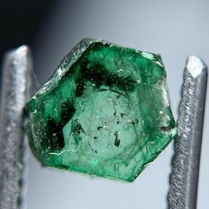Könnte beinhalten: Nahaufnahme eines rohen, sechseckigen Smaragd-Edelsteins. Der Edelstein ist ein tiefes, durchscheinendes Grün mit sichtbaren Einschlüssen und Unvollkommenheiten. Er wird von einer silbernen Metallpinzette gehalten und zeigt seinen natürlichen, unpolierten Zustand.