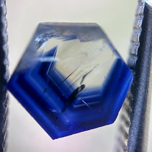 May include: A macro shot of a hexagonal gemstone with a striking blue and clear color gradient. The gem is held by metal tweezers, showcasing its intricate internal structure and facets. The gem's edges are a deep blue, transitioning to a lighter, transparent center.