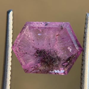 Peut inclure: Une pierre précieuse hexagonale facettée de couleur rose translucide. La gemme présente des inclusions sombres et est maintenue par une pince métallique. Les facettes de la gemme captent la lumière.