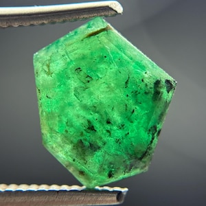 Op de afbeelding: Een levendige groene smaragd, vastgehouden door een zilveren pincet. De smaragd heeft een facetvormige, onregelmatige vorm met donkere insluitsels en een doorschijnende kwaliteit. De edelsteen wordt gepresenteerd tegen een vage grijze achtergrond.