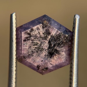Könnte beinhalten: Ein sechseckiger, facettierter Edelstein, der von einer Metallpinzette gehalten wird. Der Edelstein hat eine hellviolette Farbe mit dunklen Einschlüssen und inneren Streifen. Der Stein wird vor einem verschwommenen, neutralen Hintergrund gehalten.