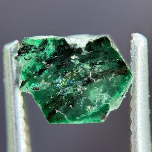 Könnte beinhalten: Ein roher Smaragdedelstein, gehalten von einer silbernen Pinzette. Der Smaragd ist ein leuchtendes Grün mit dunklen Einschlüssen und einer leicht irisierenden Oberfläche. Der Edelstein hat eine sechseckige Form.