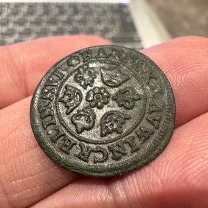 Puede incluir: Primer plano de una moneda de metal gris oscuro con un diseño circular. La moneda tiene un borde elevado y un diseño central con cuatro motivos florales estilizados. La inscripción alrededor del borde dice "CIVITATIS  HAMBVRGENSIS".