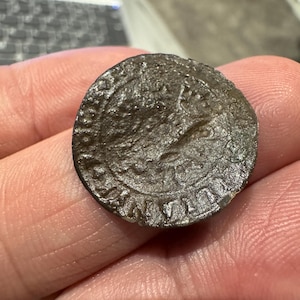 Puede incluir: Primer plano de una moneda circular de color marrón oscuro con letras en relieve alrededor del borde. La moneda tiene un aspecto desgastado y antiguo.