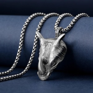 Può includere: Una collana color argento con un ciondolo a forma di testa di rinoceronte dettagliata. Il ciondolo è finemente progettato con dettagli strutturati, che mostrano le caratteristiche del rinoceronte. La catena è a maglie e lo sfondo è blu scuro.