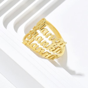 May include: A gold-colored ring with the names "Blanca", "Brian", and "Laurie" cut out in a stacked design. The ring is made of a shiny, reflective metal and sits on a white surface.