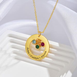 May include: A gold-colored pendant necklace featuring a circular charm with the names "John", "Caitlyn", and "Alyssa" engraved around the edge. The charm has three colored gemstones: blue, green, and red. The necklace is displayed on a white surface.