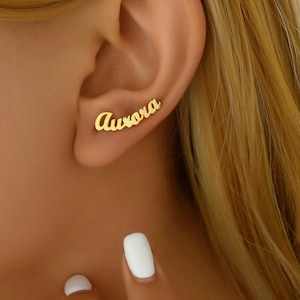May include: A gold-colored ear cuff with the name "Aurora" in a cursive font. The jewelry is worn on the ear, with the name following the curve of the ear. The background shows a person's skin and hair.