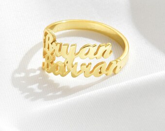 May include: A gold-colored ring with the names "Bryan" and "Barron" cut out in a cursive font. The ring is set against a white background, highlighting the jewelry's design and color. The ring is a personalized accessory.