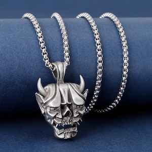 May include: Silver-toned pendant necklace featuring a detailed oni mask design. The pendant has horns, sharp teeth, and intricate facial features. The chain is a linked style, and the background is a solid blue.