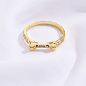 Puede incluir: Un anillo dorado con un diseño en forma de hueso que lleva el nombre "Rocky" y una huella de pata. La banda del anillo está adornada con pequeñas piedras preciosas transparentes. El anillo se muestra sobre una superficie blanca.