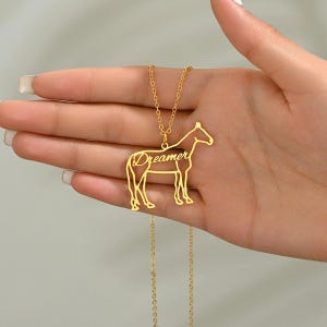 Op de afbeelding: Een goudkleurige ketting met een paardenhanger. De hanger heeft het woord "Dreamer" op het lichaam van het paard. De ketting wordt in een hand gehouden.