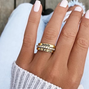 May include: Three gold rings stacked on a finger. The top ring is engraved with the name "Jacob". The middle ring is set with small, clear stones. The bottom ring displays the date "8.2.20".