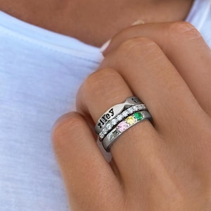 Pode incluir: Um conjunto de três anéis de prata. O anel superior é gravado com o nome "Riley". O anel do meio tem uma fileira de pedras transparentes e três pedras preciosas coloridas: rosa, amarelo e verde. Os anéis são usados em um dedo.