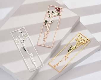 May include: Three metal bookmarks with floral designs. One is white with a snowdrop, another is rose gold with a flower and the name "Rainie", and the third is gold with a flower and the name "Jocelyn".