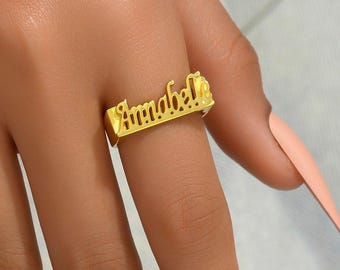 May include: A gold-colored ring with the name "Annabelle" in a cursive font. The ring is rectangular with the name cut out. The ring is on a person's finger.