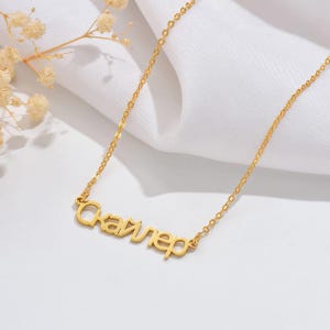 May include: A gold-colored necklace with the name "Скайлер" in Cyrillic script. The pendant is connected to a delicate chain. The necklace is displayed on a white surface with a sprig of dried flowers.