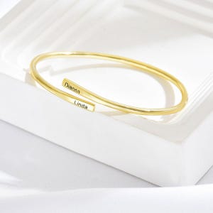 May include: A gold-coloured, open cuff bracelet. The bracelet has two rectangular bars with the names "Dianna" and "Linda" engraved on them. The bracelet is displayed on a white surface.