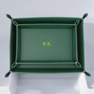 Könnte beinhalten: Ein grünes Leder-Tablett mit den Initialen "R.K." in goldfarbenen Buchstaben. Das Tablett hat eine rechteckige Form mit abgerundeten Ecken und ist für kleine Gegenstände konzipiert. Die Ränder sind hochgeklappt und mit goldfarbenen Nieten befestigt.
