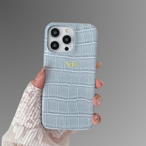May include: A pale blue phone case with a crocodile skin effect. The case features a gold monogram with the letters "NB". The case is designed for a smartphone with multiple camera lenses.